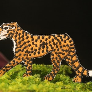 May include: An embroidered cheetah patch with a black and orange color scheme. The cheetah is walking with its head turned to the side. The patch is on a green mossy surface.