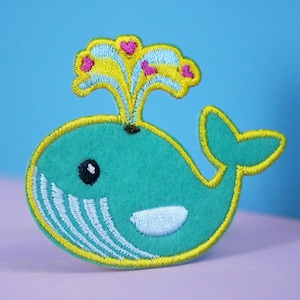 Könnte beinhalten: Ein blauer Wal mit einem gelben und weißen Wasserstrahl und rosa Herzen. Der Wal ist auf einem Filz-Patch gestickt.