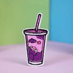 May include: Embroidered patch featuring a purple and pink drink cup with a straw. The cup design includes a planet, clouds, and a heart. The patch has a white border and is set against a colorful background.