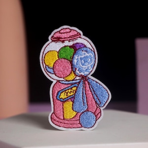 May include: Embroidered patch depicting a gumball machine. The design features a pink base, a clear globe filled with colourful gumballs, and a blue dispenser. The patch has a white border and is set against a neutral background.