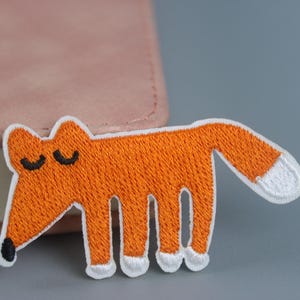 Peut inclure: Patch brodé de renard orange avec des accents blancs et des yeux noirs. Le renard est debout sur quatre pattes et a une queue recourbée.