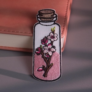 Op de afbeelding: Geborduurde patch met een doorzichtig flesontwerp met een kurk. In de fles zit een roze vloeistof en een tak met roze en witte bloemen. De patch heeft een witte rand.