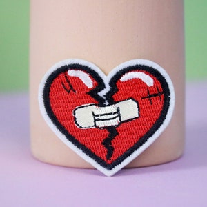 May include: A red embroidered heart patch with a white bandage over a crack. The heart is outlined in black thread.