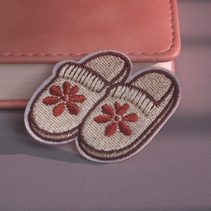 May include: An embroidered patch featuring a pair of beige slippers with dark brown trim and a red flower design on the top of each slipper. The patch has a white border and is shaped to outline the two slippers positioned side by side.