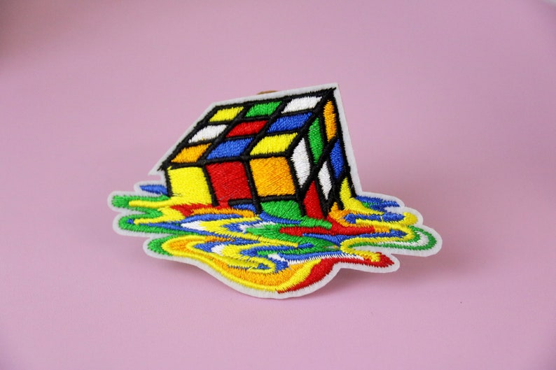 May include: A colorful embroidered patch of a melting Rubik's Cube with a white background.