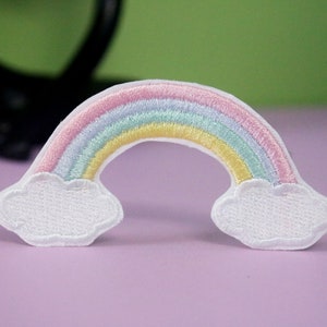 Könnte beinhalten: Ein pastellfarbener Regenbogen mit weißen Wolken an beiden Enden. Der Regenbogen besteht aus rosa, hellblau, gelbem und grünem Stickgarn.