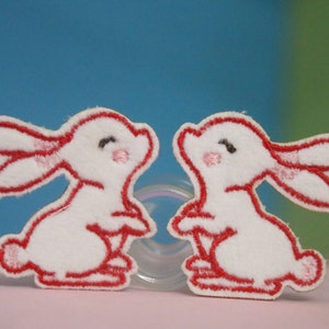May include: Two white felt bunny appliques with red outlines. The bunnies are facing each other and have pink noses and black eyes.
