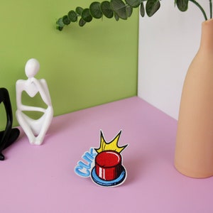 May include: A red button with a yellow star and the word "CLIK" in blue letters. The button is on a pink surface with two white and black figurines.