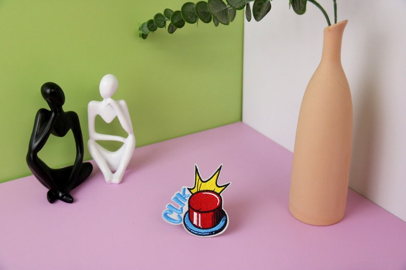 May include: A red button with a yellow star and the word "CLIK" in blue letters. The button is on a pink surface with two white and black figurines.