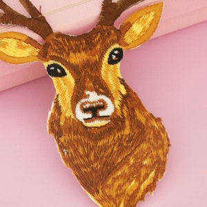 May include: Embroidered patch featuring a deer head with brown and yellow thread. The deer has large antlers, a white patch on its nose, and black eyes. The patch is likely for clothing or accessories.