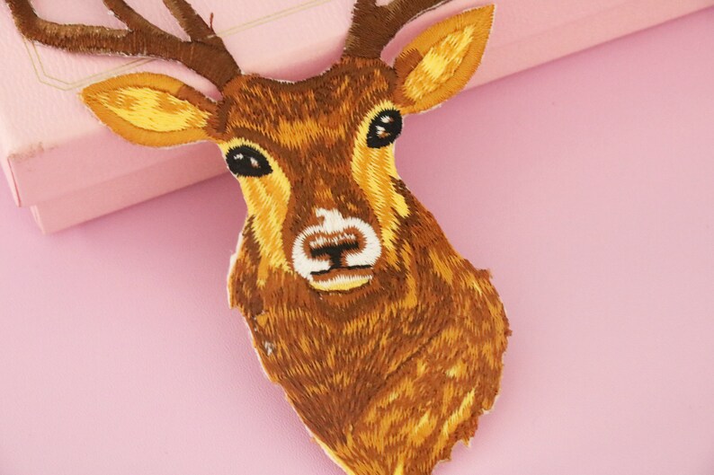 May include: Embroidered patch featuring a deer head with brown and yellow thread. The deer has large antlers, a white patch on its nose, and black eyes. The patch is likely for clothing or accessories.