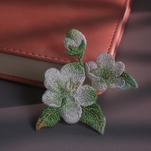 Könnte beinhalten: Ein gestickter Aufnäher mit weißen Blüten mit grüner Mitte, einer geschlossenen Blütenknospe und grünen Blättern mit goldfarbenen Garnakzenten. Das florale Design ist in detaillierten Stichen ausgeführt und zeigt weiße Blütenblätter.