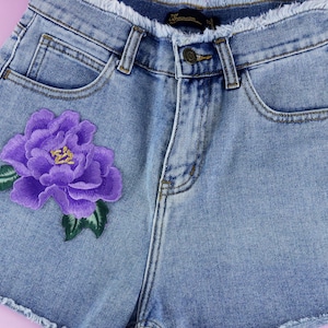 Puede incluir: Pantalones cortos de mezclilla azul claro con una flor bordada de color morado en el bolsillo delantero.