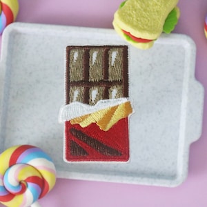 May include: A brown chocolate bar with a red wrapper and white and gold foil. The chocolate bar is on a white tray with a white background.