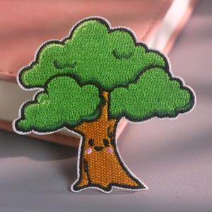 May include: An embroidered patch featuring a cartoon tree with a brown trunk and a vibrant green leafy canopy. The tree trunk has a simple face with two black eyes, a small smile, and two pink circular cheeks, all outlined in black thread.
