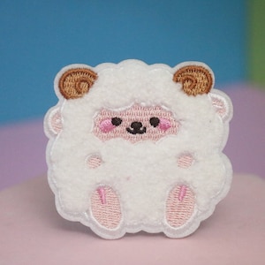 May include: White fluffy sheep applique with brown horns and pink cheeks. The sheep is embroidered with a cute face and has a fluffy texture.