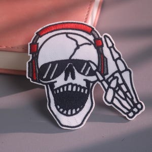 Op de afbeelding: Een geborduurde patch met een witte schedel met een open zwarte mond, die een zwarte zonnebril en een rood-zwarte koptelefoon draagt. Een skeletachtige hand is naast de schedel opgeheven, met de wijsvinger omhoog wijzend.