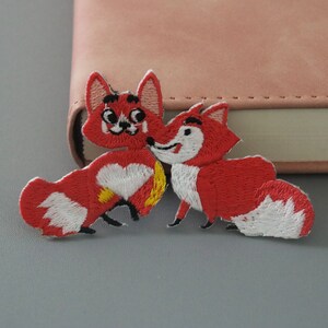 May include: Two red embroidered fox patches with white accents. The foxes are facing each other and one has a white heart on its chest.