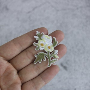 Puede incluir: Parche de flores blancas bordadas con centros amarillos y hojas verdes.