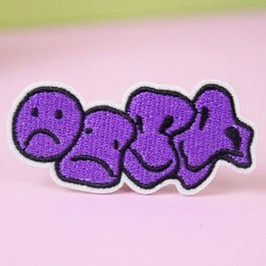 May include: Purple embroidered iron-on patch featuring four cartoon faces, each with a different expression. The faces are arranged in a row, with the first face frowning, the second face frowning, the third face smiling, and the fourth face frowning.