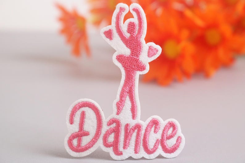 Op de afbeelding: Geborduurde patch met een roze ballerina silhouet en het woord "Dance" in cursief. De patch heeft een witte rand. De achtergrond is wazig, met oranje bloemen. Geschikt voor kleding.