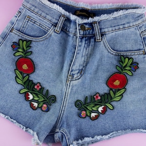 Puede incluir: Pantalones cortos de mezclilla azul claro con bordes deshilachados y diseños florales bordados en rojo, verde, blanco y azul.
