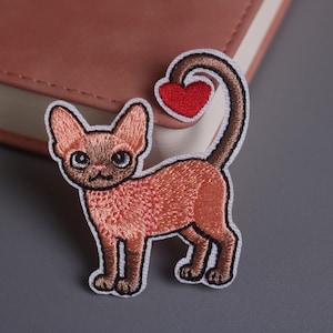 Può includere: Toppa ricamata raffigurante un gatto stilizzato con coda a forma di cuore. Il gatto è principalmente marrone con bordo bianco, e il cuore è rosso. Ideale per applicare su abbigliamento o accessori.