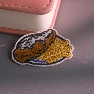 May include: An embroidered patch featuring a brown and white food item on a blue and white plate next to a pile of yellow, textured food. The patch has a white border and is shaped like a small, detailed food serving with distinct thread textures.