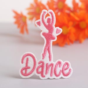 Può includere: Toppa ricamata con una ballerina rosa e la parola "Dance" in corsivo. La toppa ha un bordo bianco. Lo sfondo è sfocato, con fiori arancioni. Ideale per gli appassionati di danza.