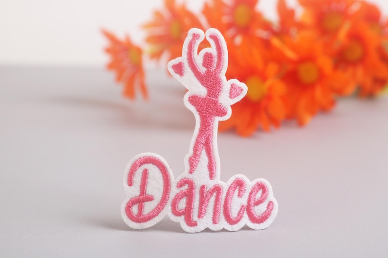 Op de afbeelding: Geborduurde patch met een roze ballerina figuur en het woord "Dance" in sierlijke letters. De patch heeft een witte rand. De achtergrond is wazig, met oranje bloemen.