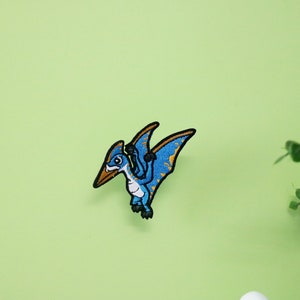 Pode incluir: Etiqueta bordada azul e amarela de um pterod&aacute;ctilo de desenho animado com um bico longo e asas abertas.