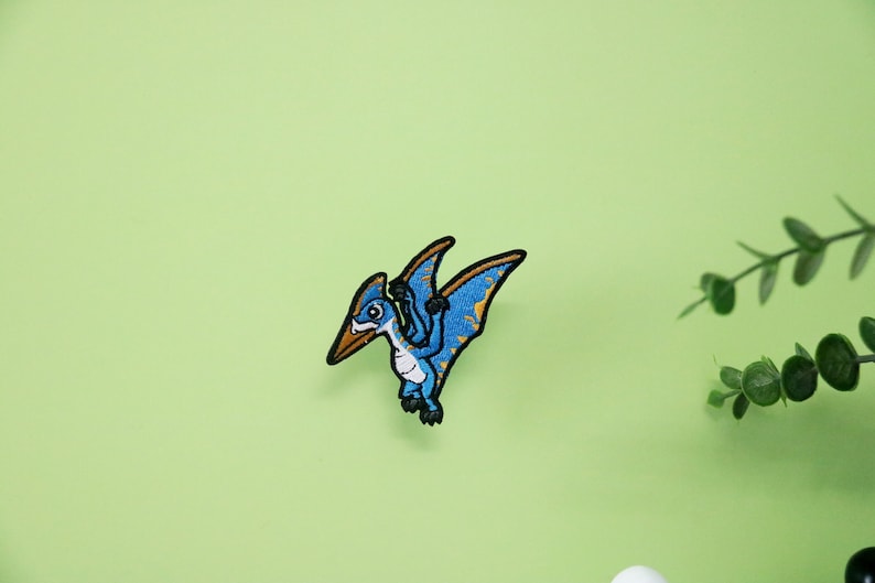 Pode incluir: Etiqueta bordada azul e amarela de um pterod&aacute;ctilo de desenho animado com um bico longo e asas abertas.