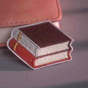 以下が含まれることがあります： 2冊の本が重なった刺繍ワッペン。上の本はダークブラウンの表紙、下の本は赤の表紙です。どちらの本も白いページで、上の本の背表紙から細い金色のしおり紐が伸びています。