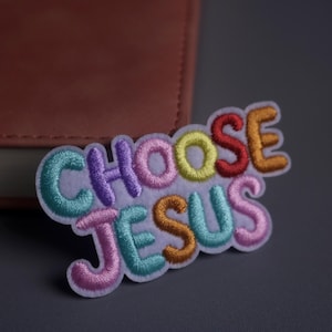 Può includere: Toppa ricamata con le parole "CHOOSE JESUS" in lettere colorate. La toppa presenta un bordo bianco ed è posta su uno sfondo grigio con un libro in pelle marrone parzialmente visibile.