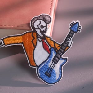 May include: An embroidered patch featuring a skeleton with a pompadour hairstyle, wearing an orange jacket, white shirt, and red tie. The skeleton is playing a blue electric guitar and making a rock and roll hand gesture with the other hand.