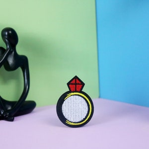 May include: A black embroidered iron-on patch with a white ring and a red diamond on top. The ring is outlined in yellow.