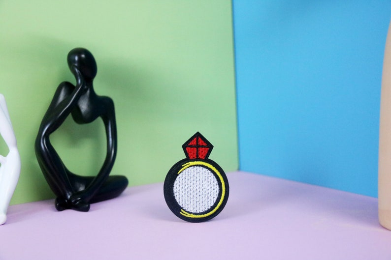 May include: A black embroidered iron-on patch with a white ring and a red diamond on top. The ring is outlined in yellow.