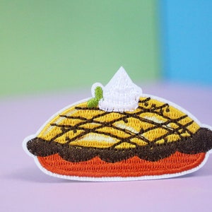 Puede incluir: Un parche bordado que representa una tarta de calabaza con una cobertura de crema batida blanca y una hoja verde, en tonos marrones y naranjas.