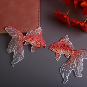 Könnte beinhalten: Zwei gestickte Goldfisch-Patches in Rot- und Weißtönen mit detaillierten Flossen und Schwänzen. Die Patches liegen auf einer grauen Oberfläche, in der Nähe eines geschlossenen Notizbuchs und orangefarbener Blumen. Die Patches sind für Kleidung oder Bastelprojekte.