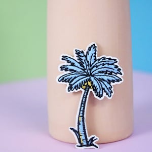 Puede incluir: Un parche de palmera azul bordado para planchar. La palmera tiene un tronco amarillo y hojas verdes.