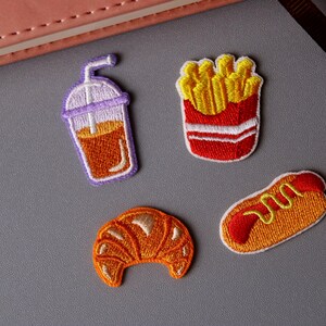 May include: Four embroidered iron-on patches featuring a croissant, a hot dog, a cup of iced coffee, and a box of french fries.