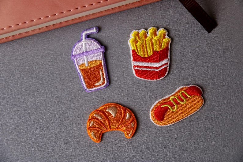 May include: Four embroidered iron-on patches featuring a croissant, a hot dog, a cup of iced coffee, and a box of french fries.