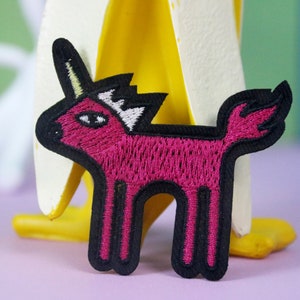 May include: Pink embroidered unicorn patch with a black outline and a white horn. The patch is on a yellow and white surface.