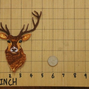 May include: Embroidered patch of a deer head with large antlers. The patch is brown and tan, with detailed stitching. A quarter is next to the patch for size comparison. The patch is approximately 3 inches tall.