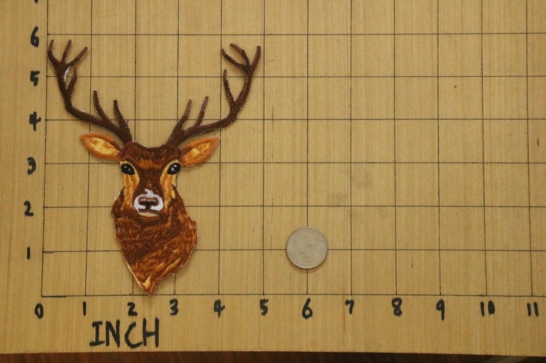 May include: Embroidered patch of a deer head with large antlers. The patch is brown and tan, with detailed stitching. A quarter is next to the patch for size comparison. The patch is approximately 3 inches tall.