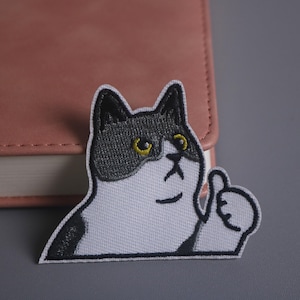 May include: Embroidered patch featuring a cat with black and white markings, yellow eyes, and a raised thumb. The patch is on a white background with a black border. The cat is giving a thumbs up.