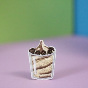 Puede incluir: Un parche bordado que representa un batido con nata montada y chispas de chocolate, en marrón y blanco.