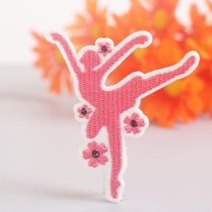 Può includere: Una toppa ricamata rosa a forma di ballerina in posa di danza. La toppa è decorata con piccoli fiori rosa con centri scuri. Lo sfondo presenta fiori arancioni sfocati.