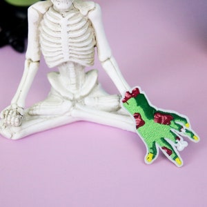 May include: A green zombie hand with red flesh and yellow fingernails is embroidered on a white fabric patch. The patch is sitting on a pink surface next to a white skeleton figurine.