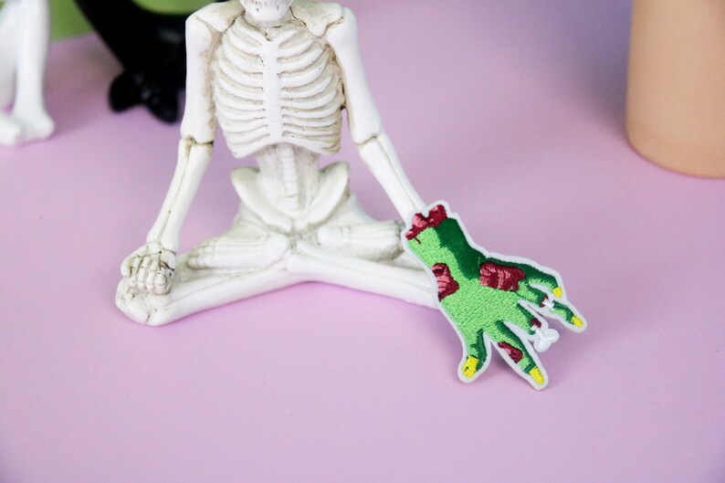 May include: A green zombie hand with red flesh and yellow fingernails is embroidered on a white fabric patch. The patch is sitting on a pink surface next to a white skeleton figurine.
